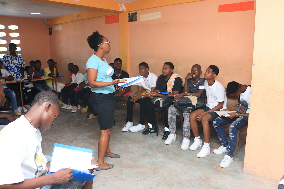 A person stands addressing a group of seated individuals in a classroom setting, holding papers and appearing to lead a session.