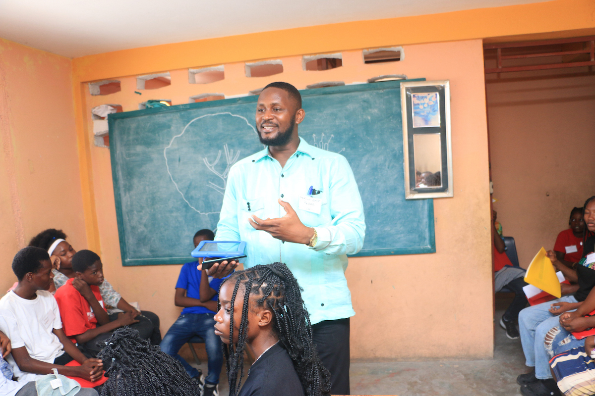 A person in a light blue shirt stands before a group of seated individuals in a classroom, holding a tablet and gesturing while speaking.