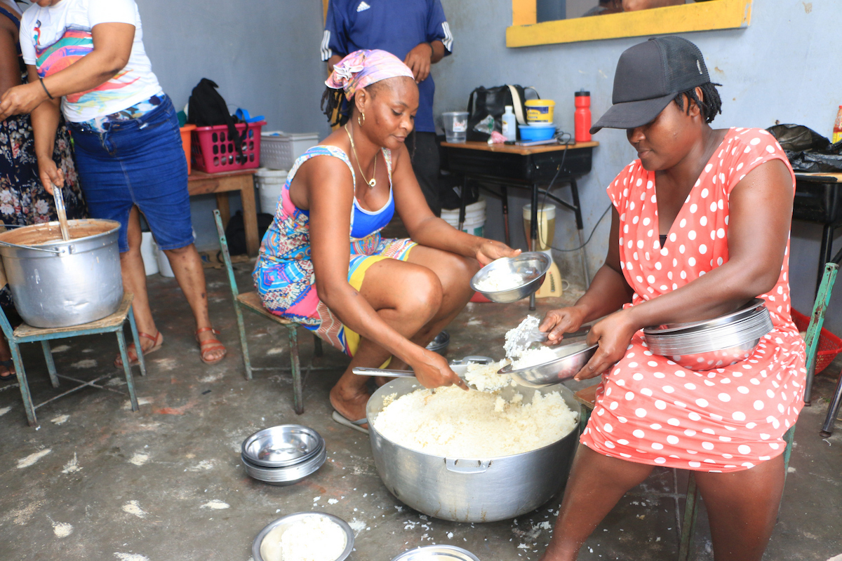 Two individuals kneel beside a large pot of rice, serving portions into metal bowls while others assist in the background.