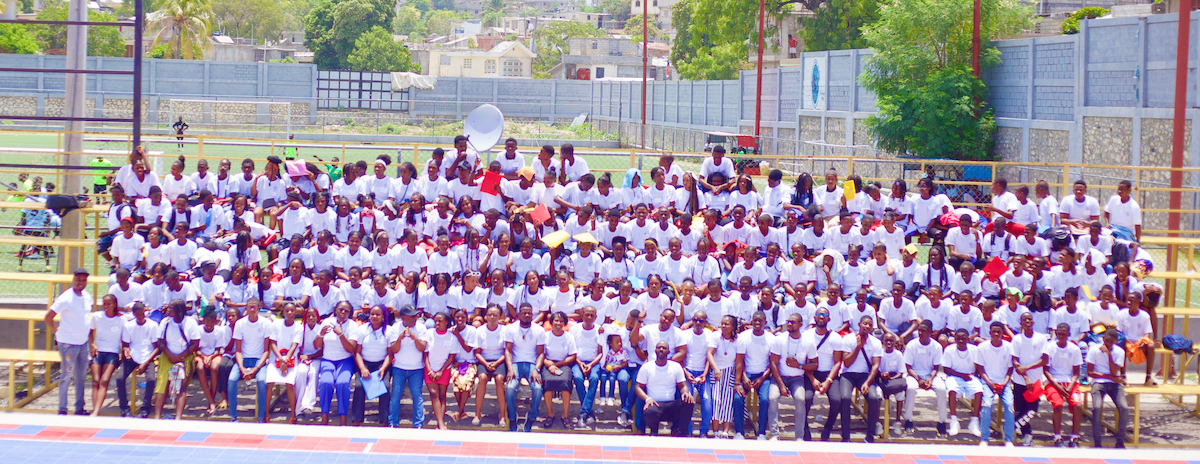 A large group of people wearing white shirts gather outdoors for a group photo on a sports field.