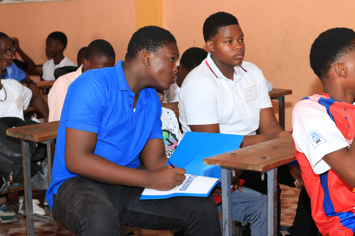 Students sit at wooden desks in a classroom, attentively listening while one writes in a blue folder.