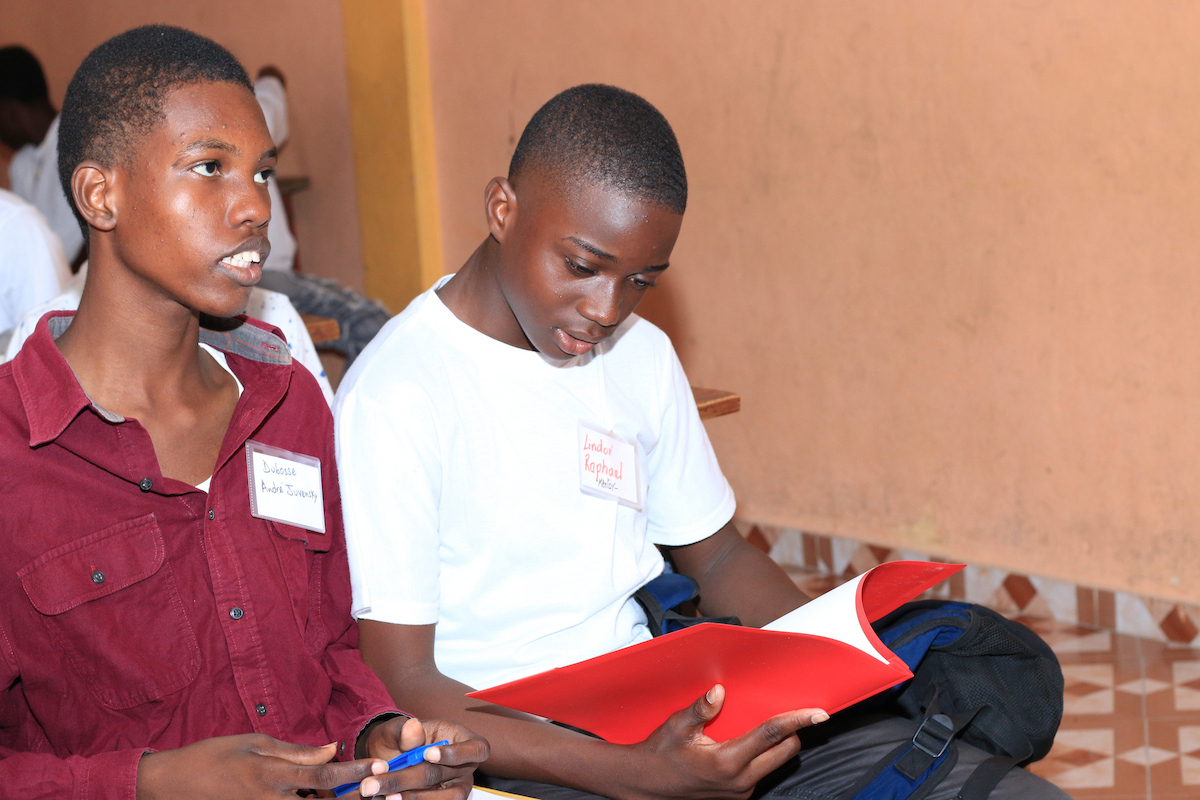 Two young individuals sit together, one holding a red folder and looking down at it while the other looks up and to the side.