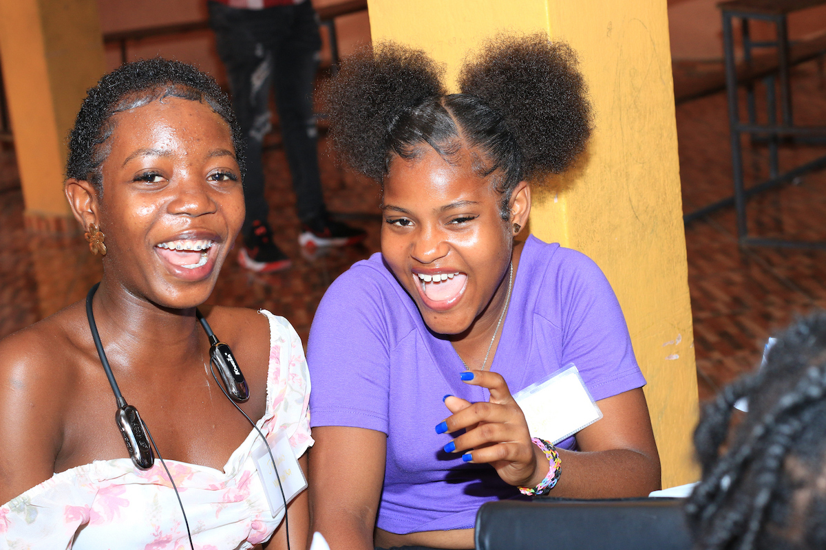 Two individuals laugh joyfully at an event, one with short hair and a floral top, the other with large puffy hair buns and a purple shirt.