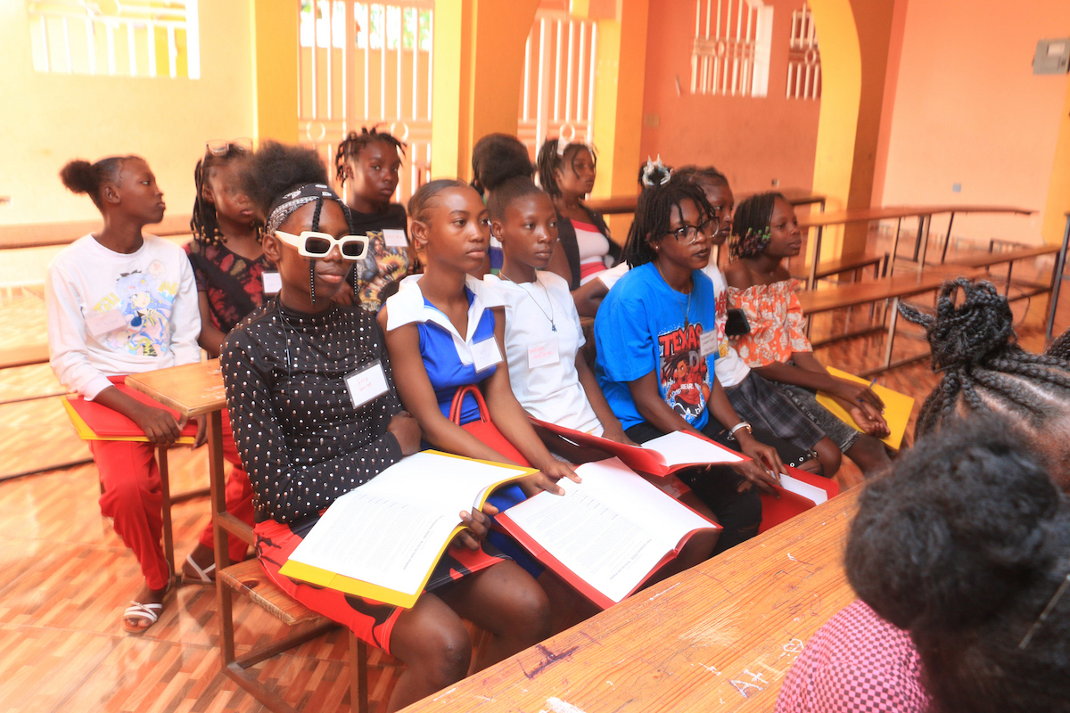 A group of young people sit attentively in a classroom, holding open books and looking toward a speaker.