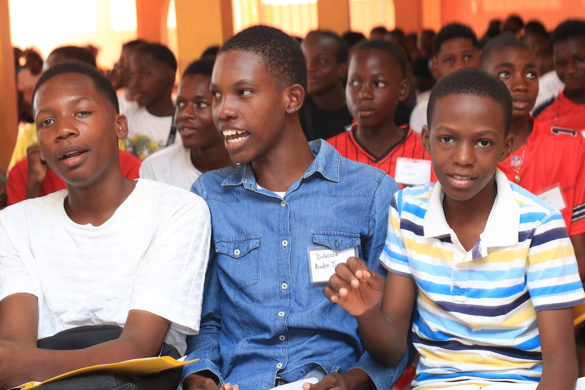 Three young people sit together in a crowd, with one smiling and gesturing while others look on attentively.