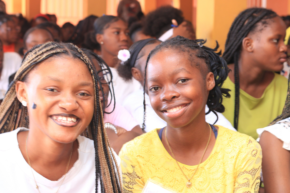 Two individuals with braided hair smile warmly at the camera while seated in a group of people.